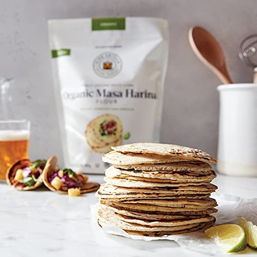 Stack of tortillas with Organic Masa Harina packaging, a glass of beer, and tacos in the background.