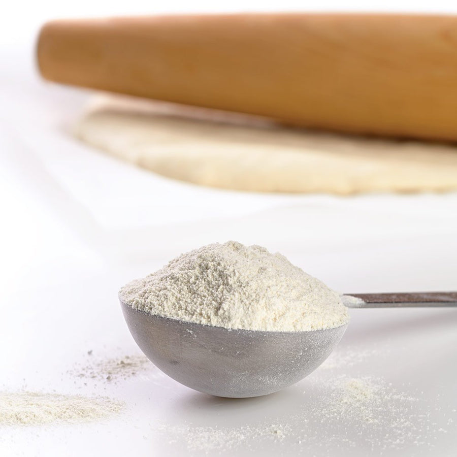 Spoonful of flour with a wooden rolling pin in the background on a white surface