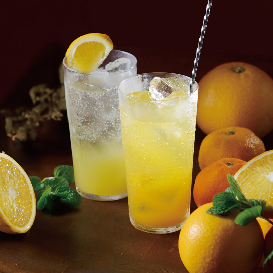 Two glasses of Yuzu juice with ice and lemon slices, surrounded by oranges and mint leaves on a wooden surface.