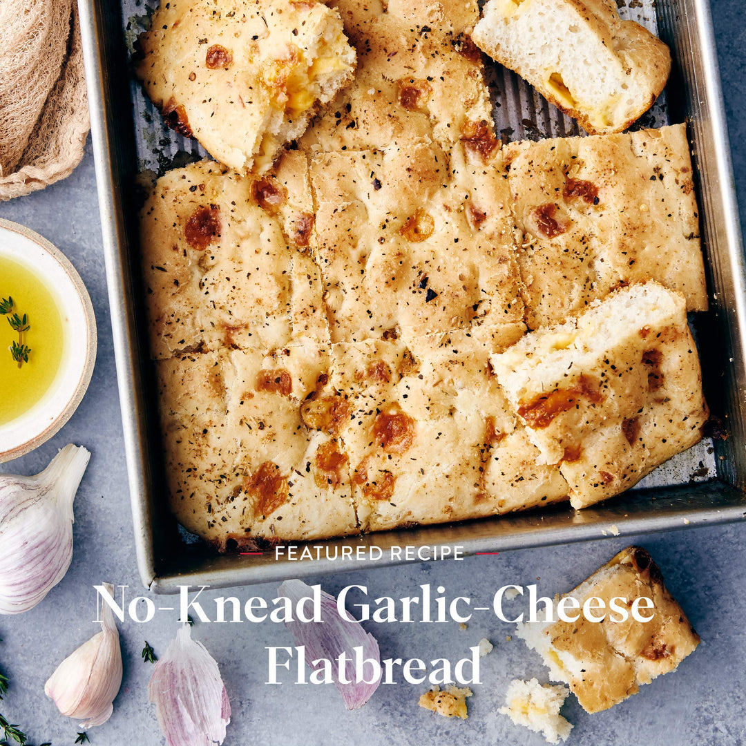 No-Knead Garlic-Cheese Flatbread in a baking tray with garlic and olive oil on a gray surface.