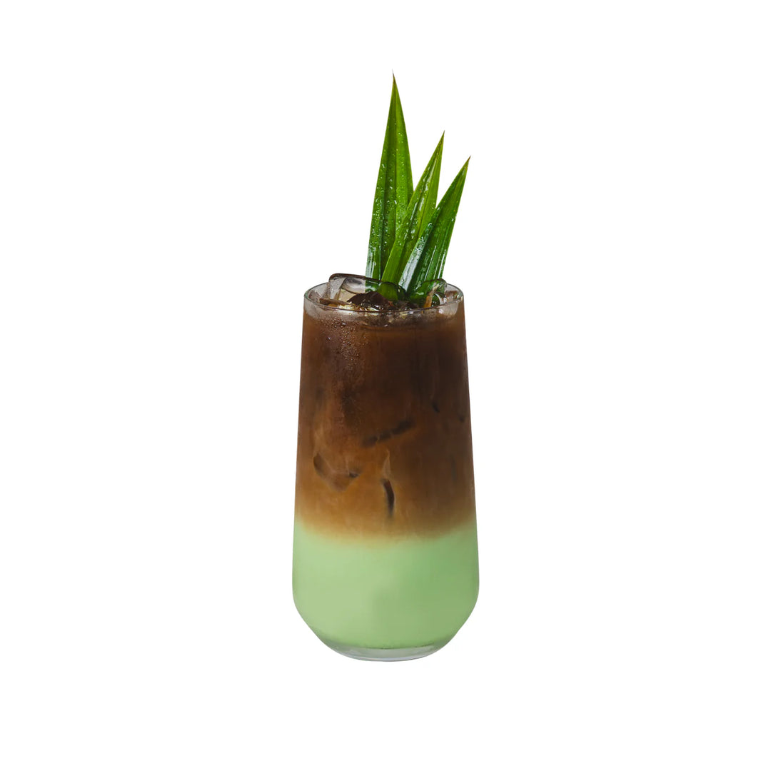 Two-toned drink in a glass with a green bottom with pandan powder and brown top, garnished with a leaf on a white background. Bossen - Fun Foods canada