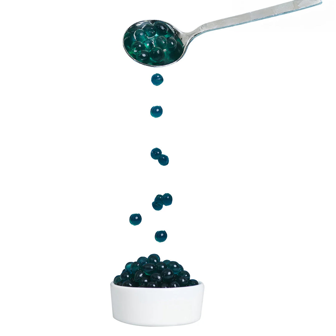 Spoon pouring blue raspberry boba capsules into a white bowl on a white background