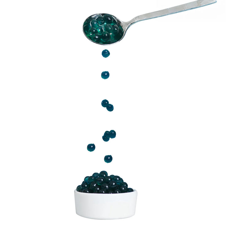 Spoon pouring blue raspberry boba capsules into a white bowl on a white background