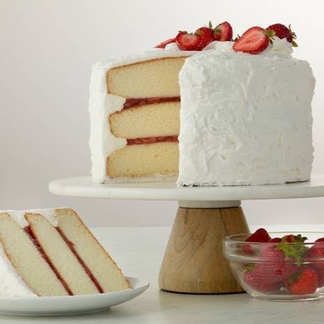 Layered cake with white frosting and strawberries on a marble stand, with a slice on a plate and additional strawberries in a bowl made with Dawn Icing Canada.