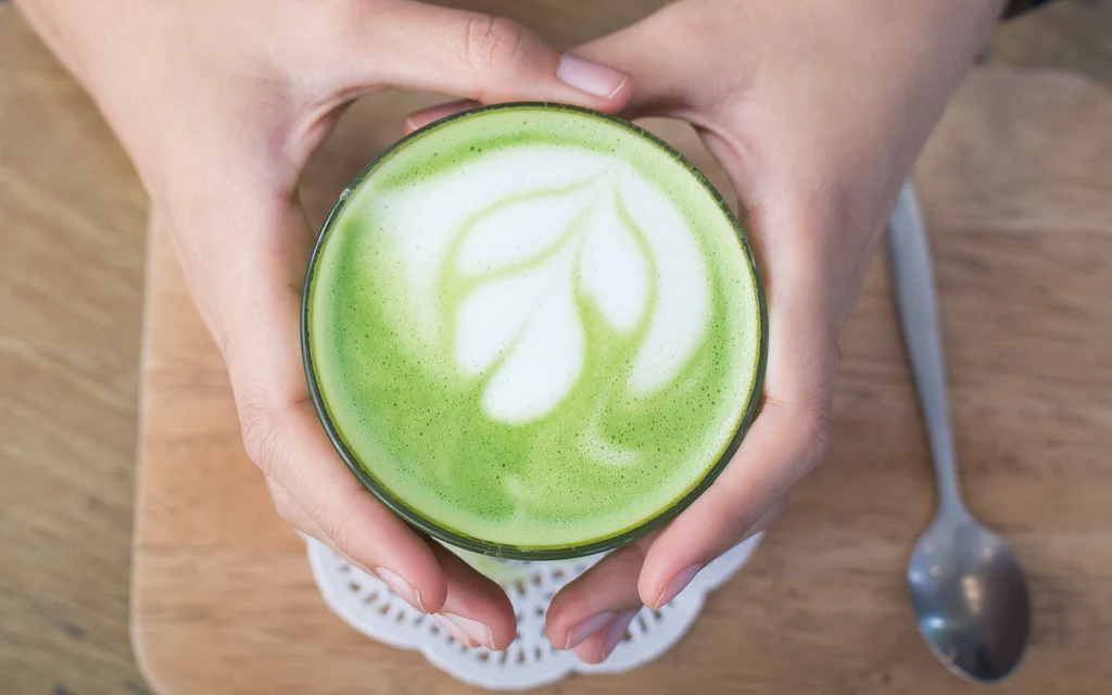 Hands holding a green matcha latte with a leaf design on a wooden table.