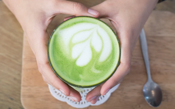 Hands holding a green matcha latte with a leaf design on a wooden table.