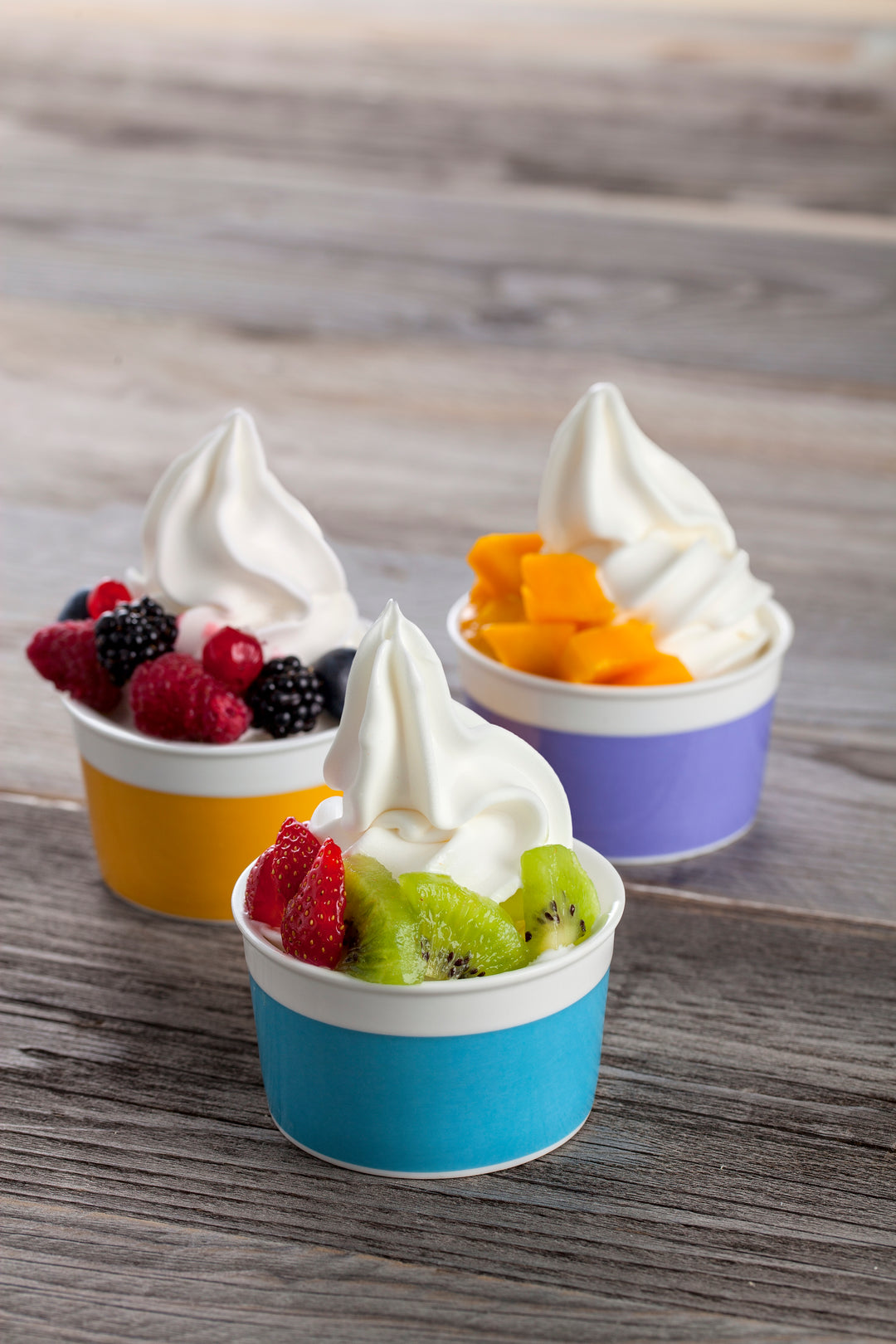 Three small cups of greek yogurt soft serve ice cream with fruit toppings on a wooden surface.