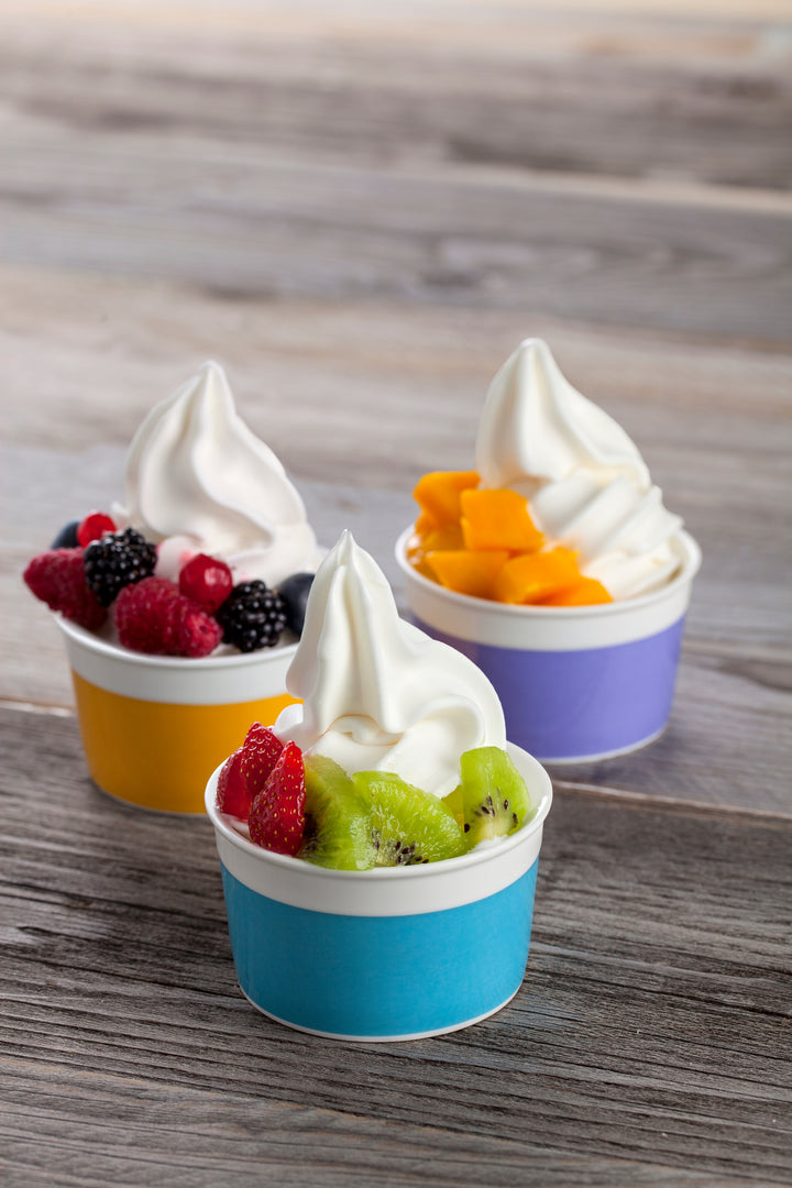 Three small cups of greek yogurt soft serve ice cream with fruit toppings on a wooden surface.
