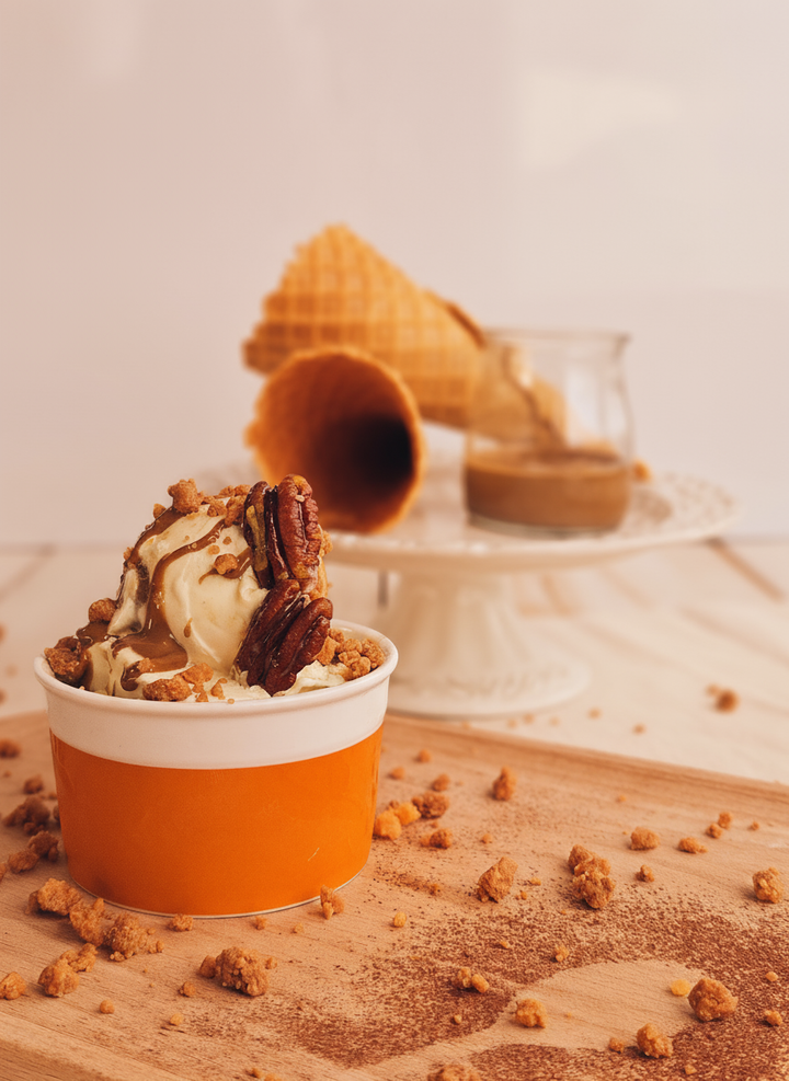 Ice cream with pecans and rubicone caramel dry crumble in an orange cup on a wooden surface with waffle cones in the background.