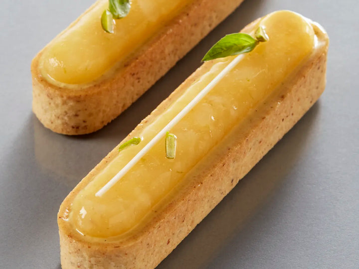 Two pastries with yellow glaze, Pure Pistachio Paste and green leaves on a gray surface.